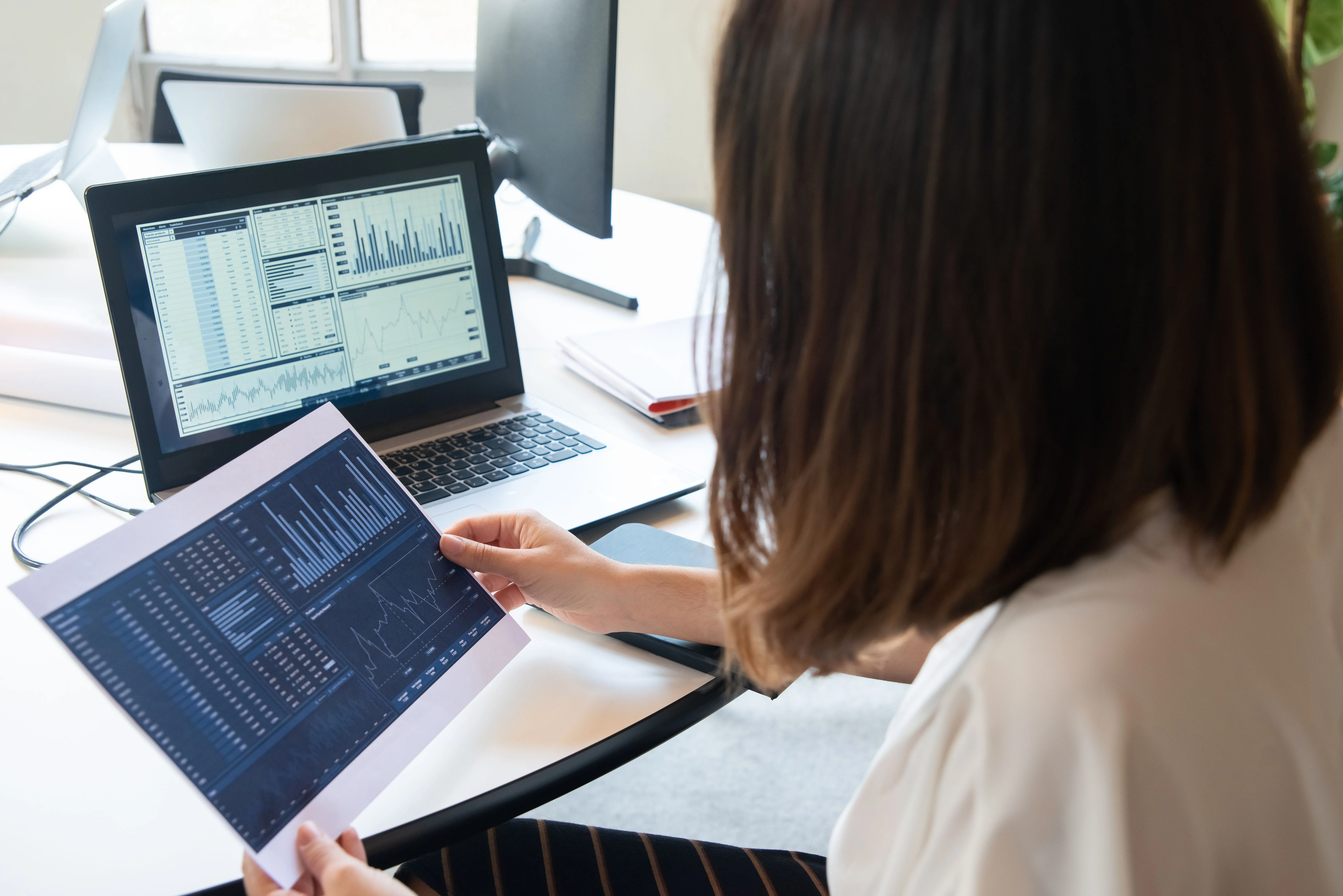 A woman with brown hair views financial data and charts displayed on a tablet and laptop at a desk, with multiple monitors visible in the background of a bright office workspace.