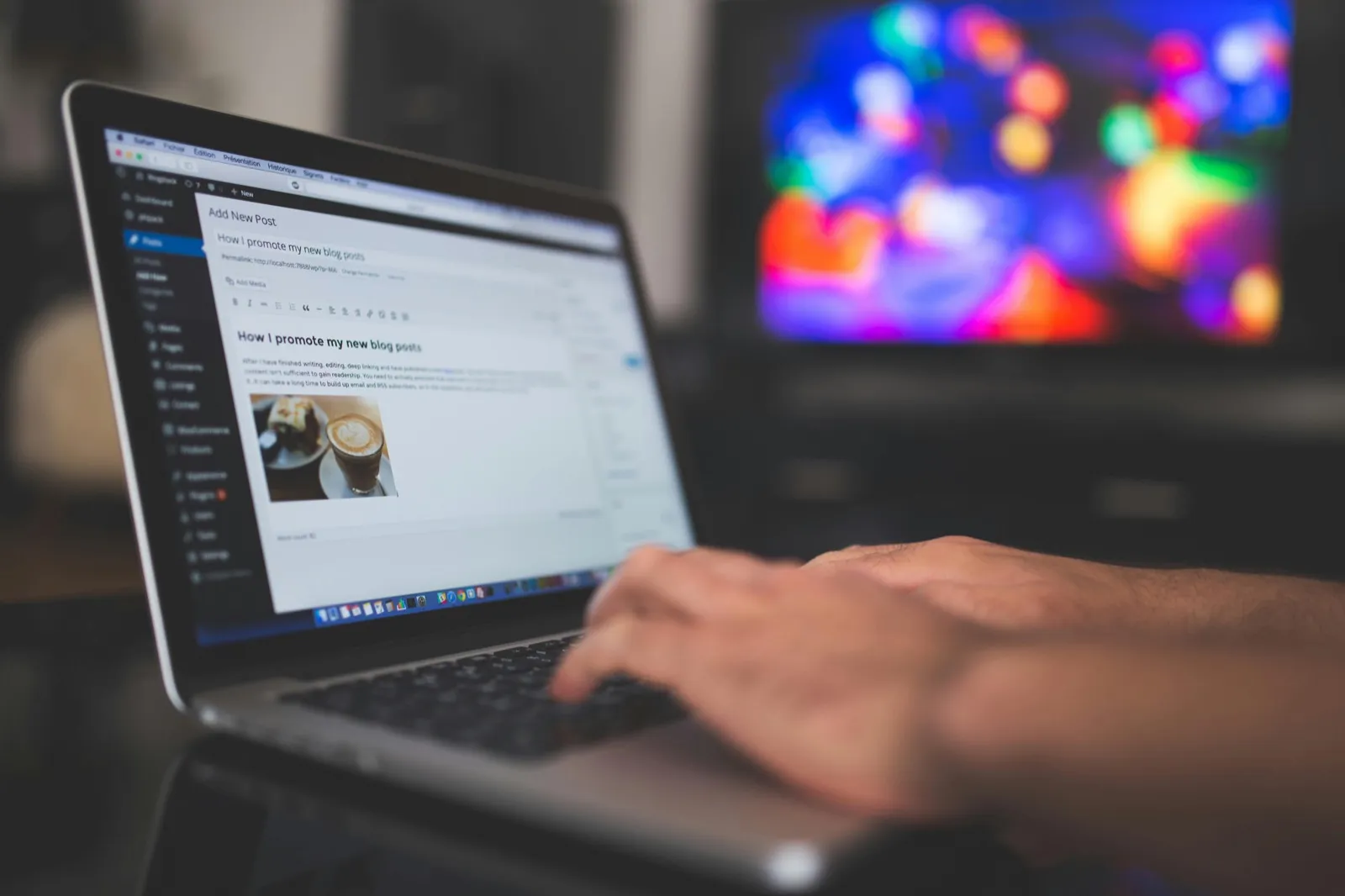 Person's hand on laptop keyboard displaying WordPress blog post editor with title "How I promote my new blog posts" and a coffee cup thumbnail, with blurred colorful lights visible on the monitor in the background.
